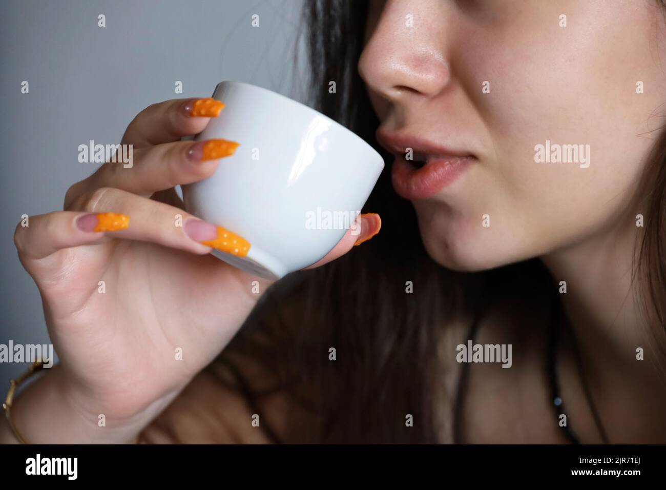 Chica sensual bebiendo té o café, taza blanca en la mano con uñas acrílicas anaranjadas cerca de los labios femeninos. Concepto de disfrutar de una bebida caliente Foto de stock