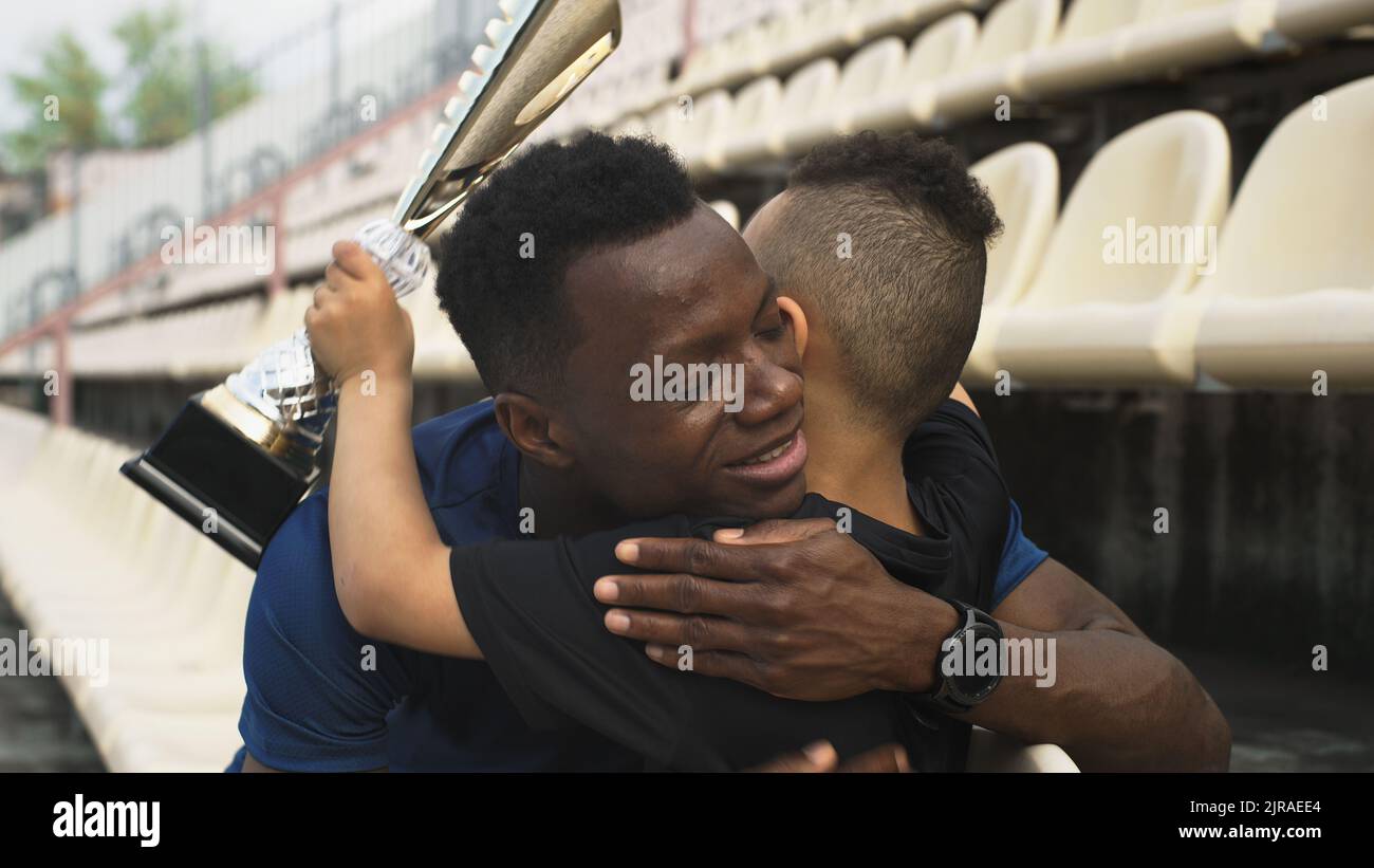 Hombre afroamericano dando copa y abrazando al chico mixto de carreras mientras estaba sentado en el estadio después de la victoria en el partido Foto de stock