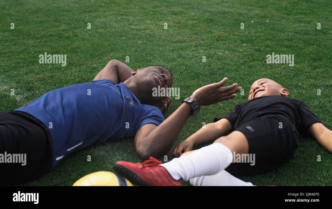 Recorra la vista del hombre negro haciendo cosquillas al chico de carreras mixto y apuntando al cielo mientras está tumbado en el campo de fútbol durante el descanso en el entrenamiento Foto de stock