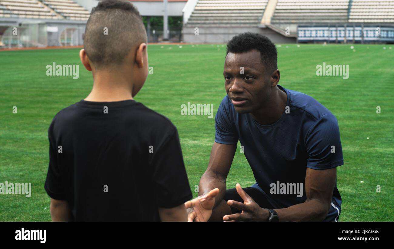 Alejar la vista del hombre negro hablando con un niño de carreras mixto practicando con el balón durante el entrenamiento de fútbol en el estadio Foto de stock