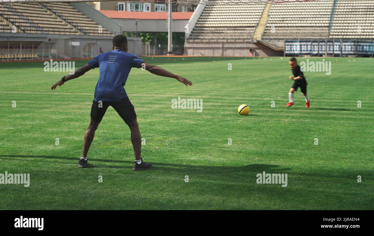 Hombre afroamericano tratando de atrapar el balón tiro de un niño mixto de carreras mientras juega al fútbol en el campo verde en el estadio en verano Foto de stock