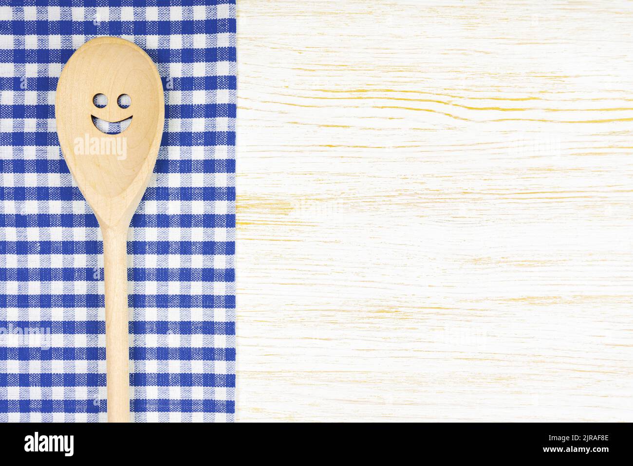 Cuchara de madera con sonrisa sobre mantel azul a cuadros sobre superficie de madera blanca. Mockup para menú o receta, restaurante, sitio web con cocina. Cocina foo Foto de stock