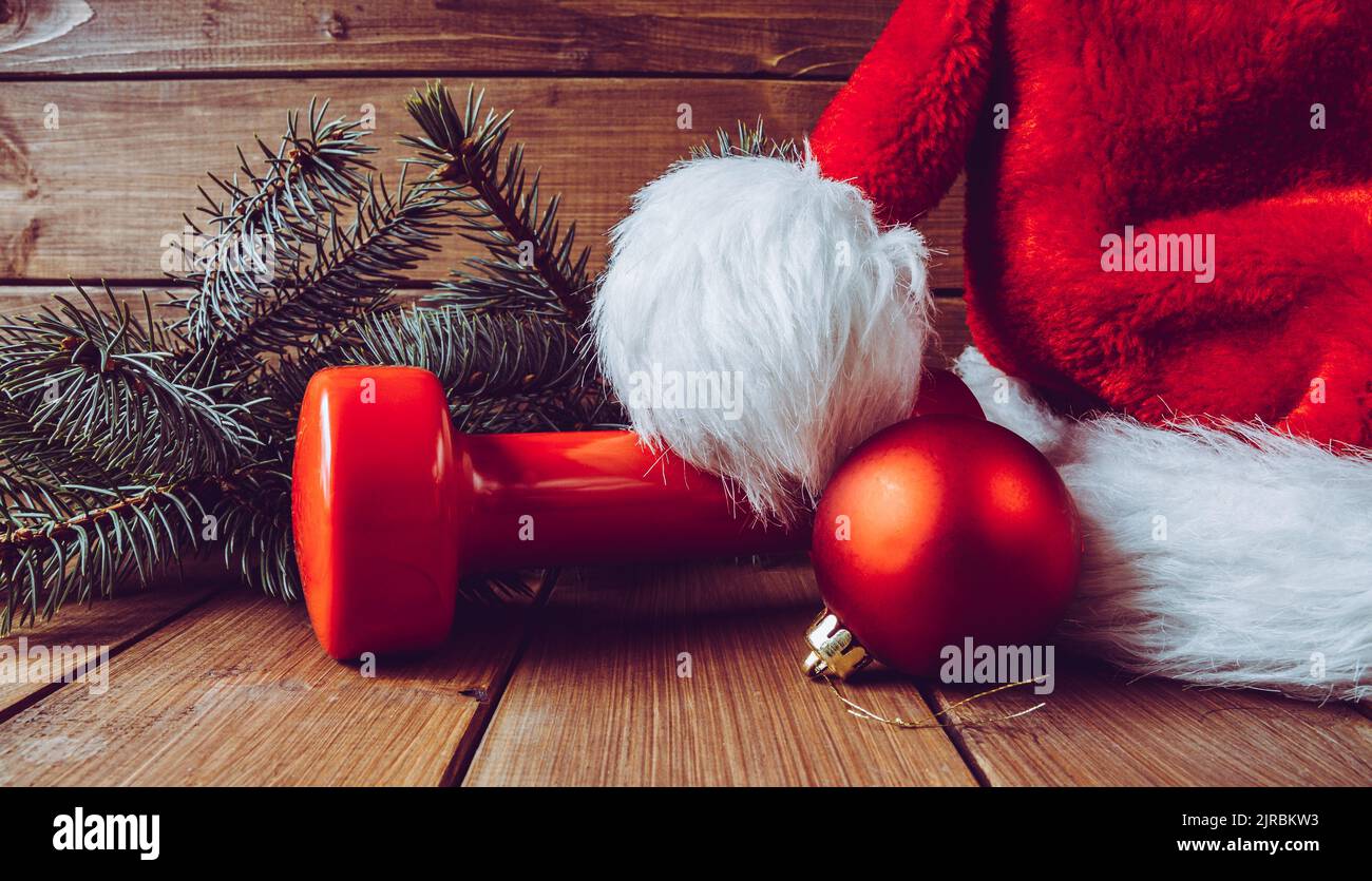 Mancuerna roja con gorro de Papá Noel. El equipo del ejercicio como idea del regalo de Navidad. Gimnasio fitness temporada concepto composición con ramas de árboles. Foto de stock