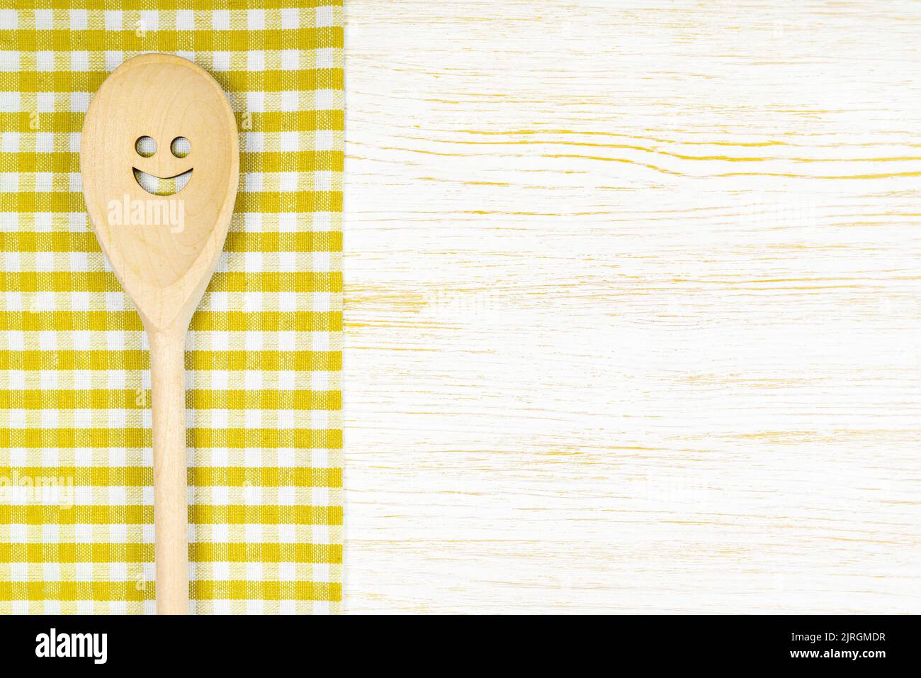 Cuchara de madera con sonrisa sobre mantel amarillo a cuadros sobre superficie de madera blanca. Mockup para menú o receta, restaurante, sitio web con cocina. Cocina f Foto de stock