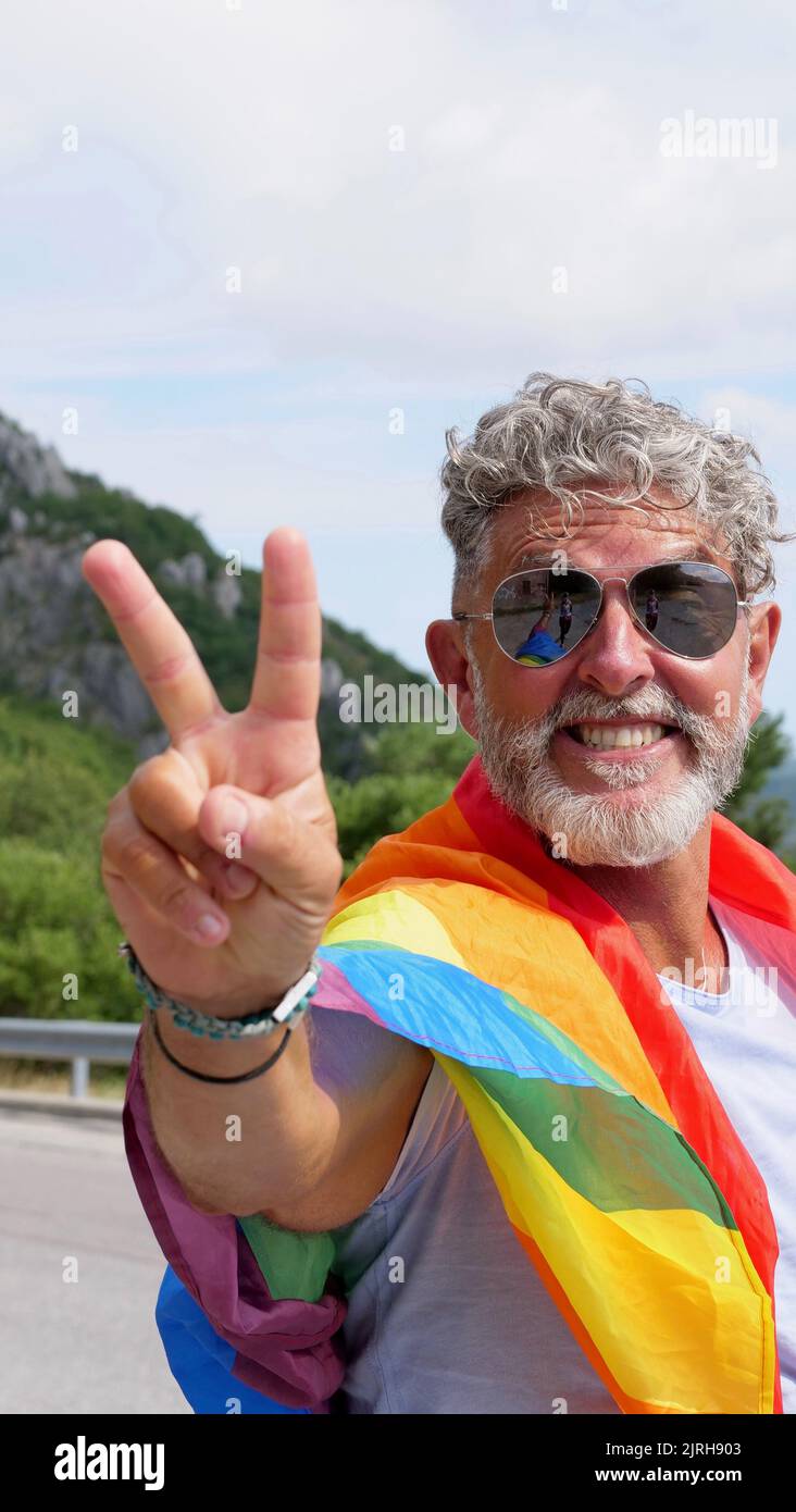 Retrato de un hombre de raza gris, anciano, caucásico, bisexualidad con barba y gafas de sol con una bandera arcoiris LGBT y un gesto de mano de paz en las montañas. Celebra el Mes del Orgullo, Día de la Bandera del Arco Iris Foto de stock