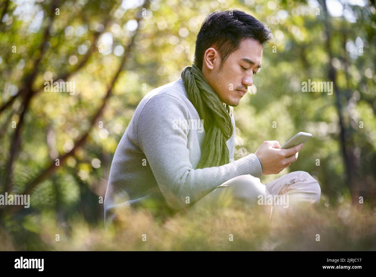 joven hombre asiático adulto sentado en la hierba al aire libre en el parque leyendo contenidos en el teléfono móvil Foto de stock