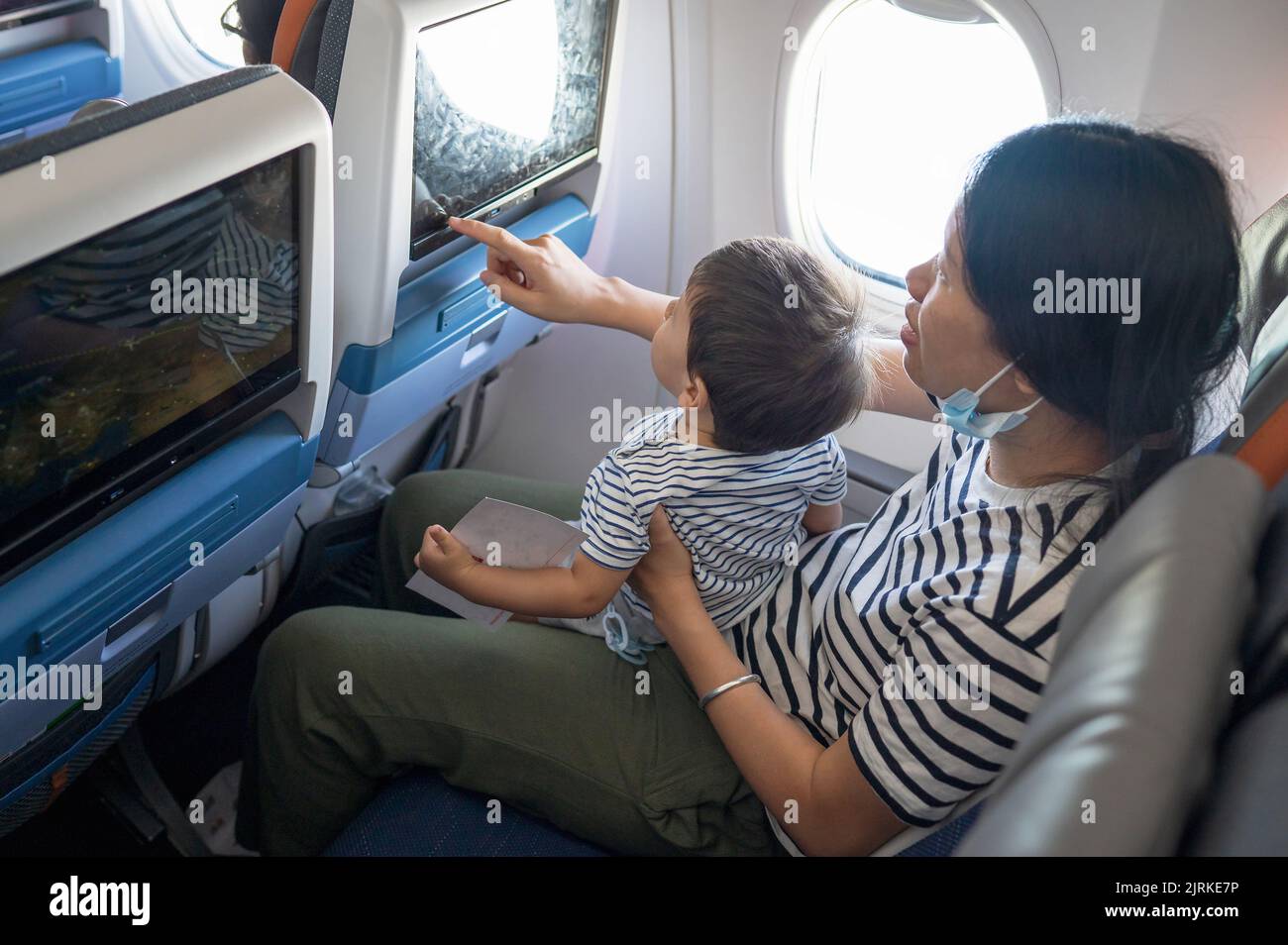 Bebé que viaja en avión sentado en su regazo con la pantalla de entretenimiento en el vuelo. Un niño de un año volando en avión y tratando de ser Foto de stock
