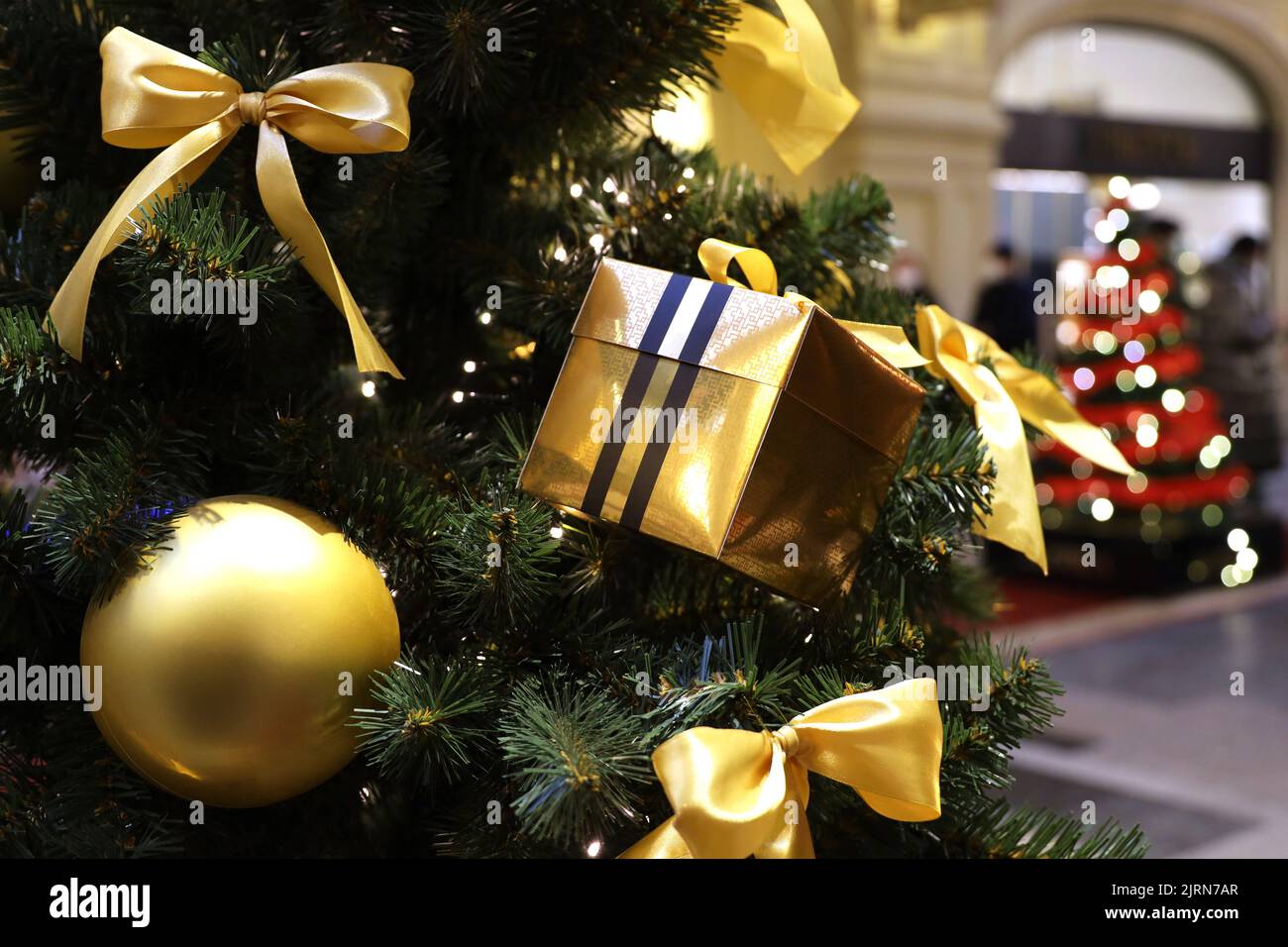 Árbol de Navidad con juguetes dorados y caja de regalo en un centro comercial sobre el fondo de luces festivas borrosas. Decoración de Año Nuevo, vacaciones de invierno Foto de stock