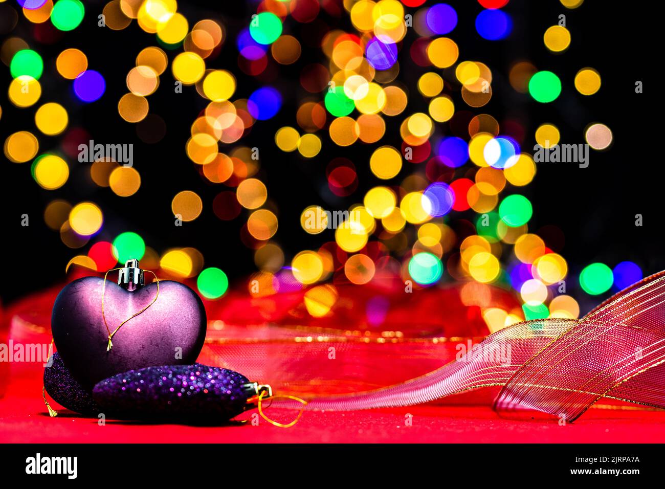 Un primer plano de ornamentos púrpura en forma de corazón para el árbol de Navidad con una cinta en un fondo de luces borrosas Foto de stock