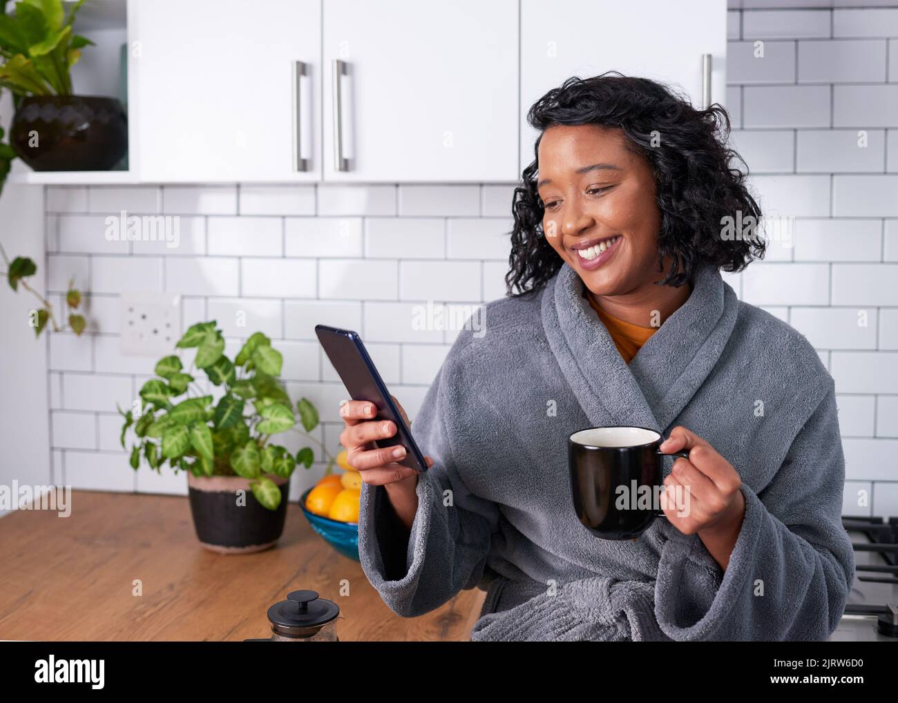 Una mujer joven sonríe mientras lee su teléfono con una bata de invierno bebiendo café Foto de stock