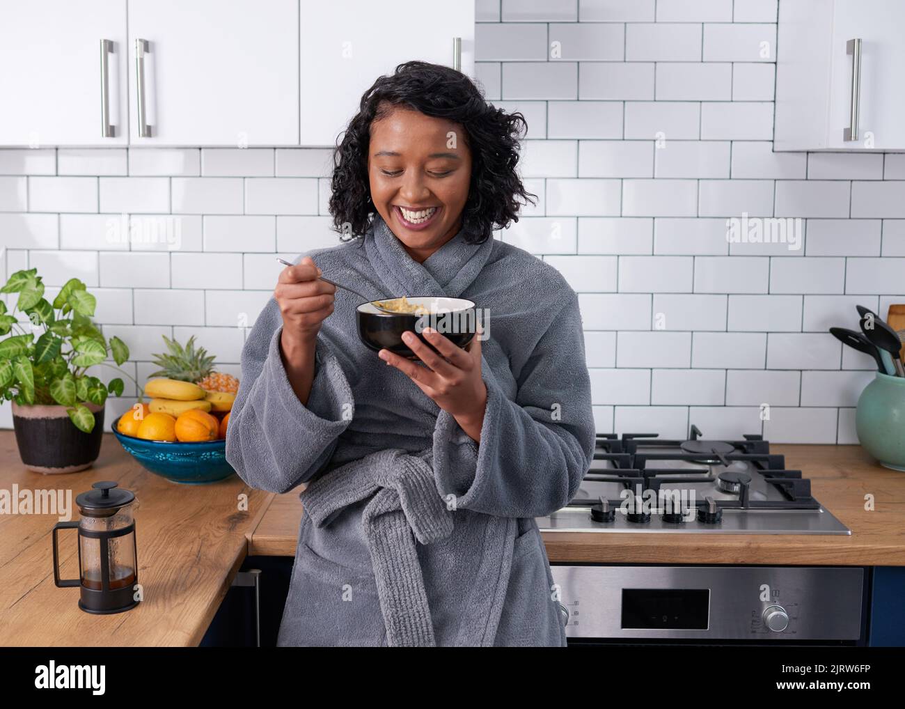 Una joven mujer multiétnica se ríe mientras come cereal de desayuno en bata Foto de stock