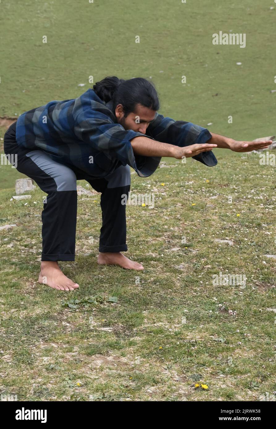 Un hombre joven indio de pelo largo que practica la postura del oso (estiramiento delantero de la variación de la postura de la silla) en al aire libre. Un joven haciendo Utkatasana yoga pose (Variation Forward Stretch). Foto de stock