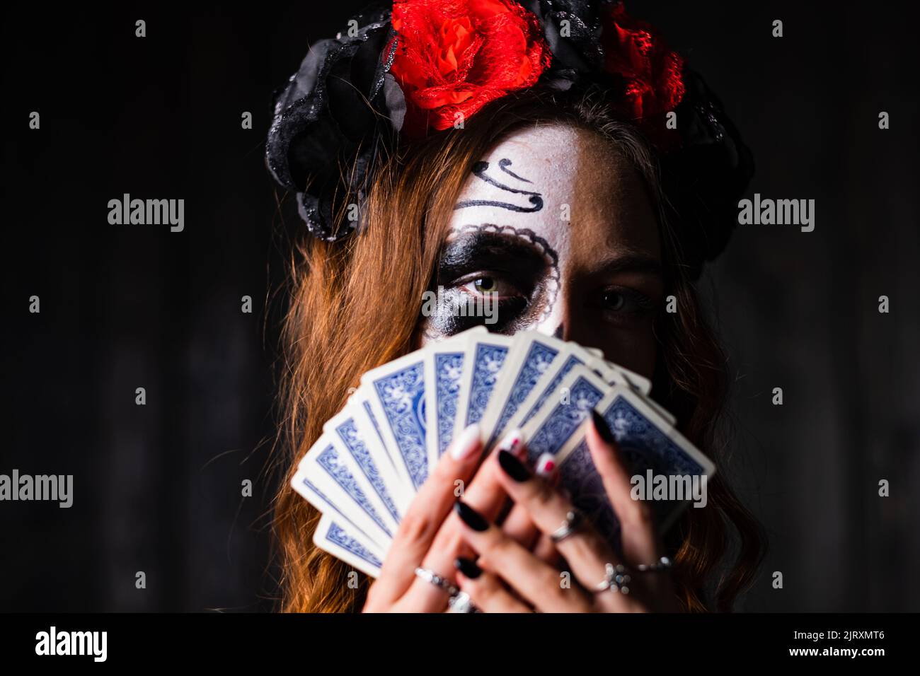 Una mujer con el maquillaje de Halloween en su cara en una corona de rosas rojas y negras se cubre con jugar a las cartas sobre un fondo oscuro Foto de stock