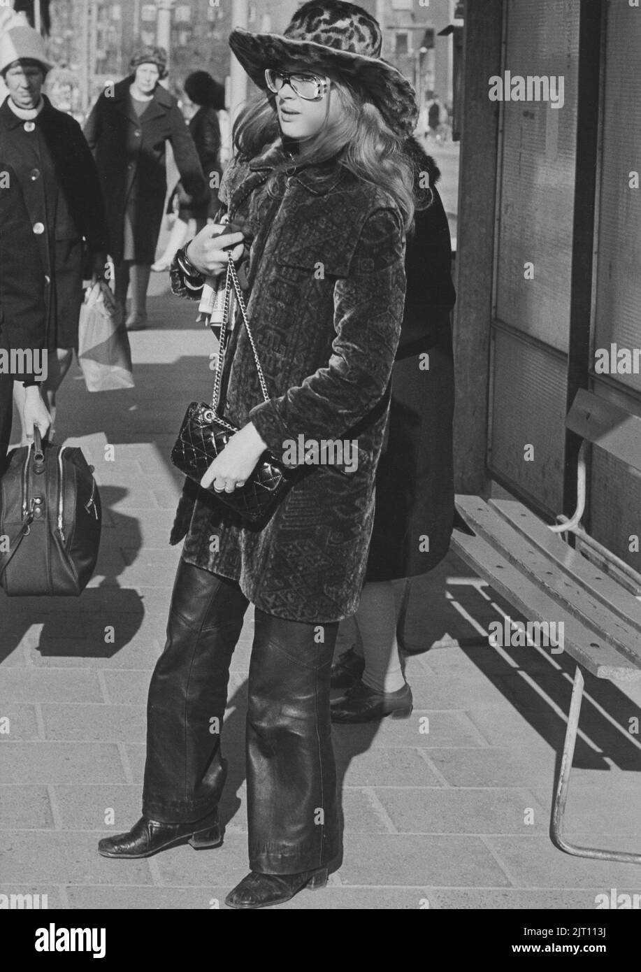 En la década de 1970s. Una mujer joven en la calle vestida con pantalones de cuero, abrigo medio largo de tela de terciopelo y sombrero a juego. Las gafas son típicas 1970s. El año es 1970. Foto de stock