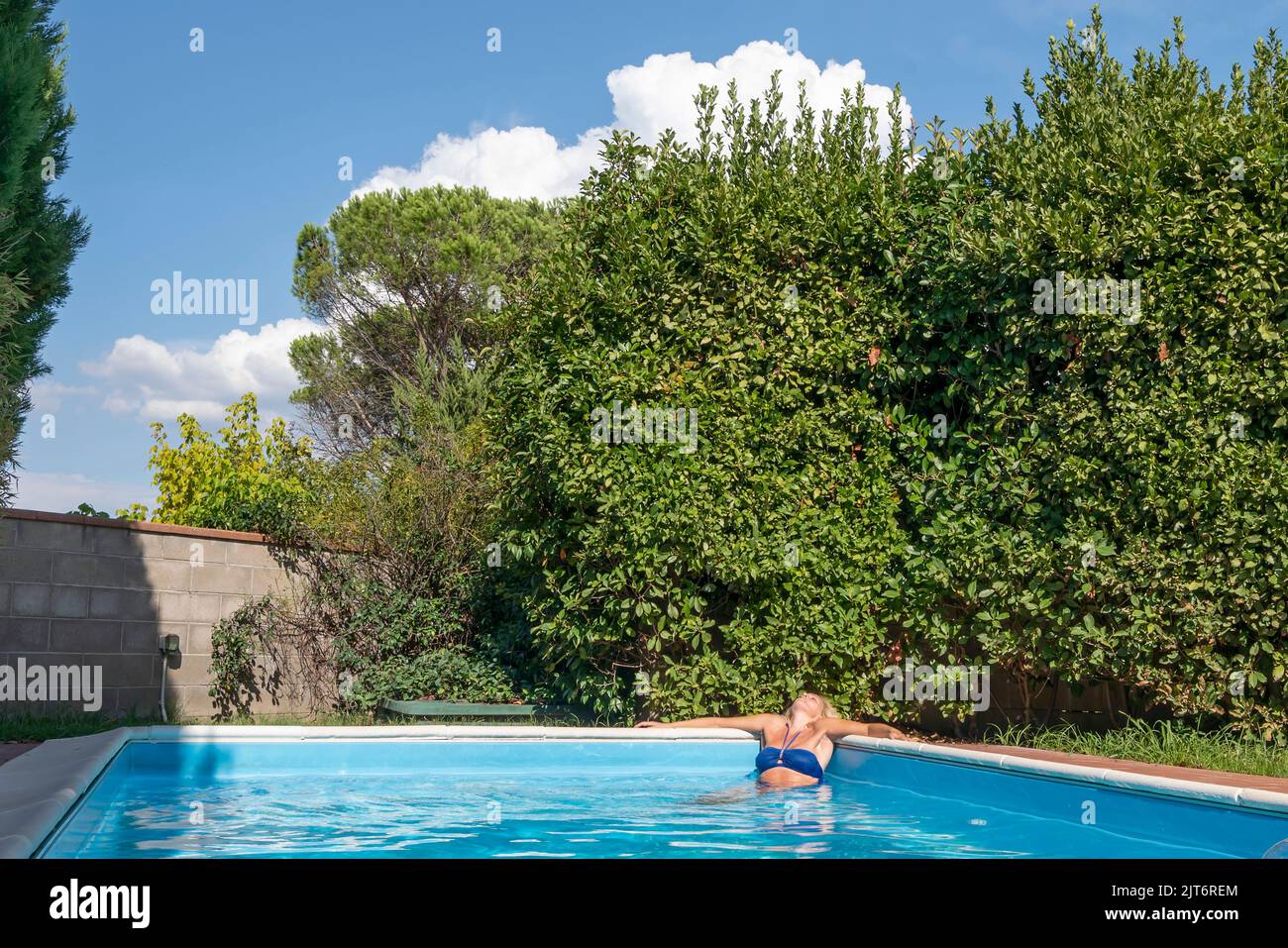 Una mujer rubia se relaja en la esquina de una piscina en un jardín con muchas plantas Foto de stock