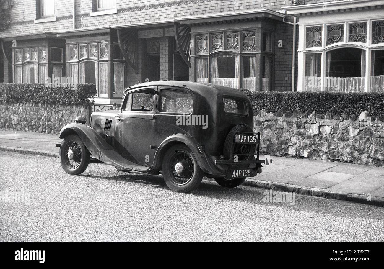 1950s, histórico, vista lateral de un pre-guerra de dos puertas Morris ocho coches de motor con ruedas de alambre, puertas de autobuses y rueda de repuesto fuera en la parte trasera, estacionado en una calle de casas adosadas, con paneles decorativos en la parte superior de la ventana, Inglaterra, Reino Unido. En la parte trasera del vehículo, dos placas de matrícula traseras, una en el parachoques y la otra en una placa de remolque. Producido por primera vez en 1935, los ocho como se conocía comúnmente, fue el coche británico más popular de finales de los años 1930s y estuvo en producción hasta 1948. Su éxito vio a Morris Motors convertirse en el mayor fabricante de motores de Gran Bretaña. Foto de stock