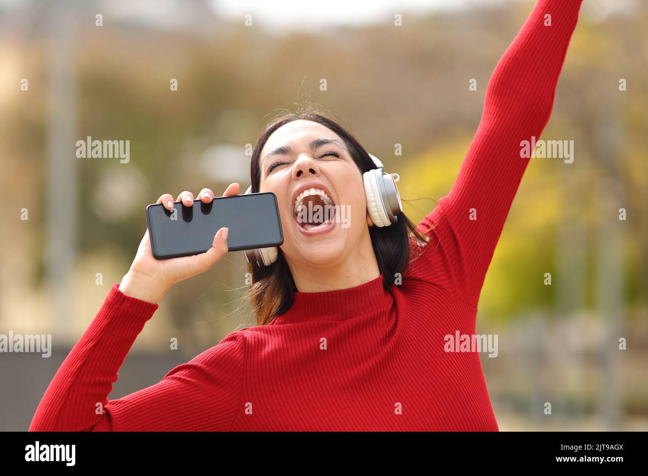Feliz mujer en rojo sosteniendo el teléfono como un micrófono cantando en la calle escuchando música con auriculares Foto de stock