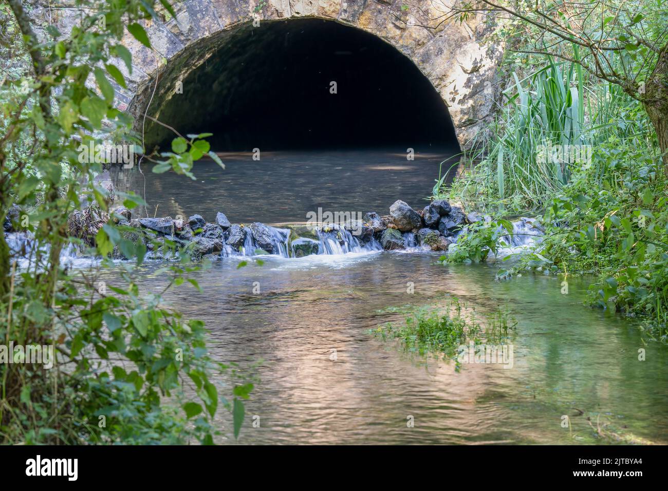 primer plano de un río de tiza que fluye fuera de un túnel de arco de piedra y sobre una pequeña cascada de piedra Foto de stock