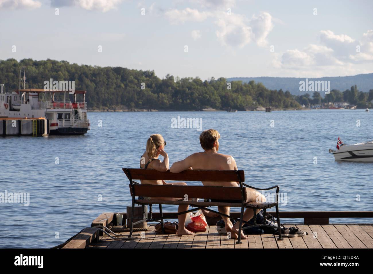 Una joven pareja disfruta del sol de la tarde mientras se sienta en un banco en el puerto de Oslo con vistas al fiordo de Oslo, Noruega, Escandinavia. Foto de stock