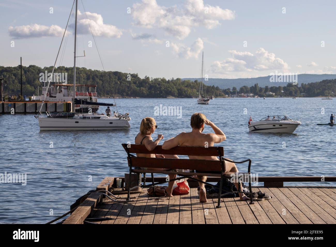 Una joven pareja disfruta del sol de la tarde mientras se sienta en un banco en el puerto de Oslo con vistas al fiordo de Oslo, Noruega, Escandinavia. Foto de stock