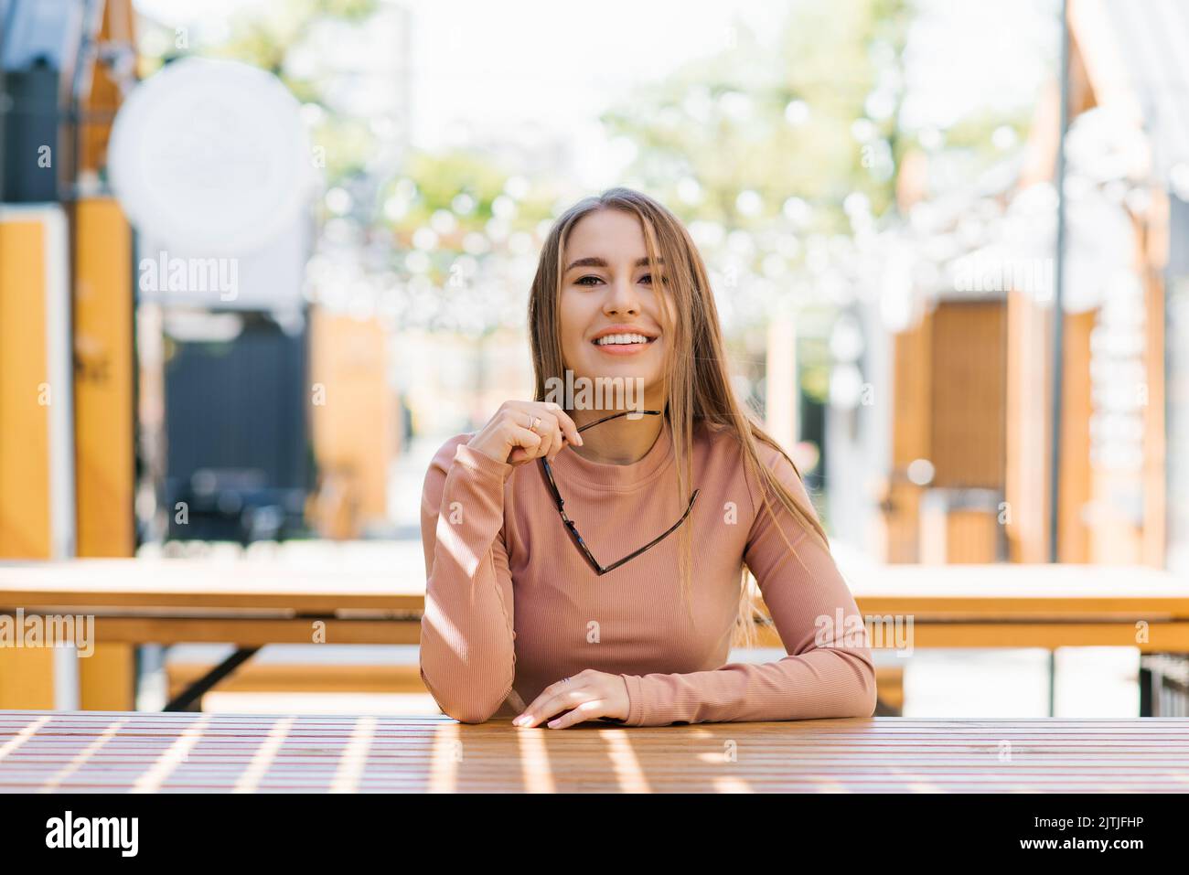 Retrato de una mujer sonriente feliz sosteniendo gafas de sol en la ciudad en verano en una cafetería y esperando un pedido Foto de stock