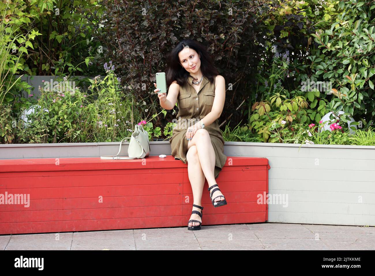 Mujer bonita con vestido hace un selfie con cámara de smartphone mientras se sienta en un banco de la calle sobre flores de fondo Foto de stock