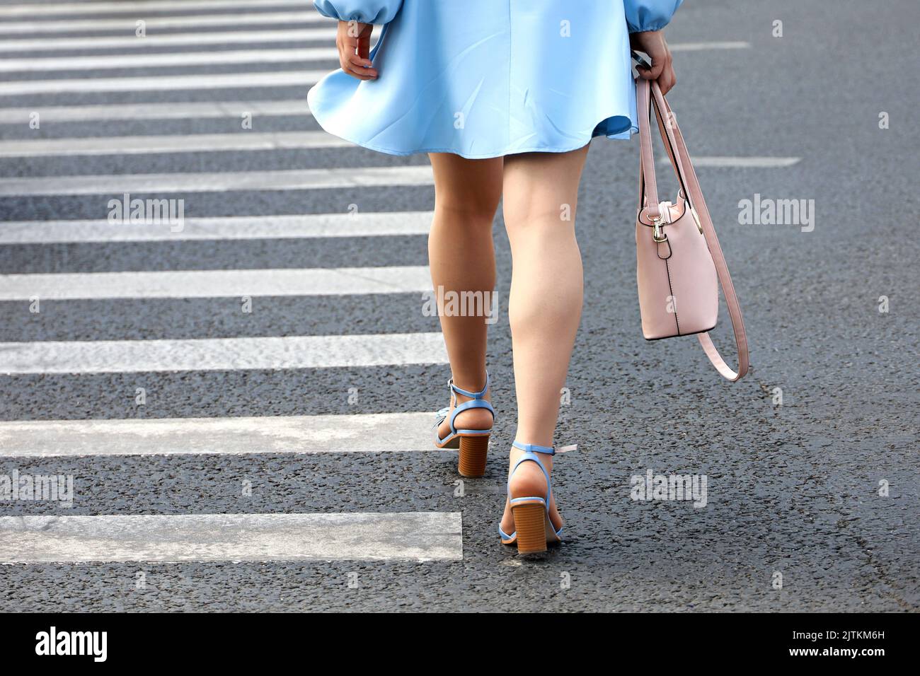 Chica con vestido corto azul y zapatos en tacones altos cruzando con bolso rosa la calle en el cruce. Piernas femeninas en el cruce de peatones Foto de stock
