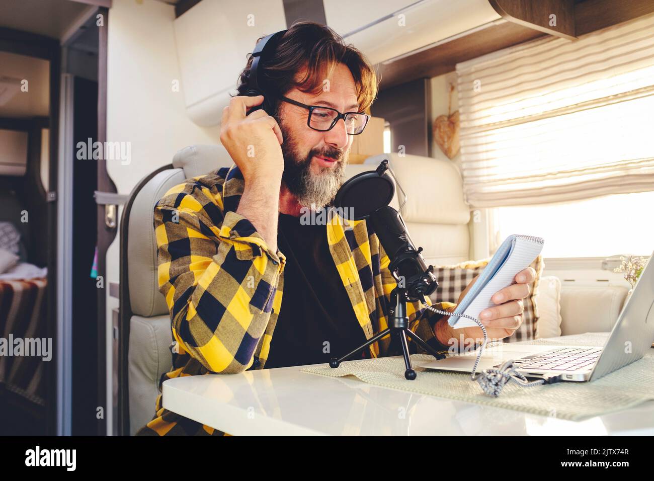 Retrato de un hombre joven y maduro con barba y gafas grabando contenido en línea con un micrófono en una casa de oficina alternativa de furgoneta camper. Vanlife Foto de stock