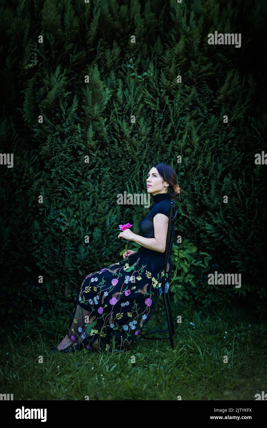 Una joven mujer bonita con ropa negra se sienta en una silla en el jardín Foto de stock