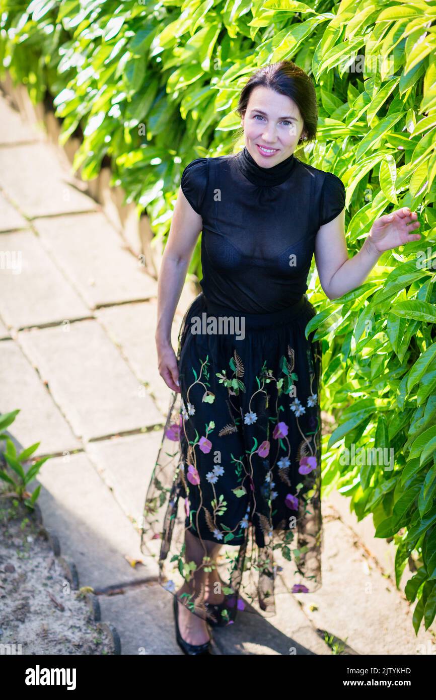 Una joven mujer bonita con ropa negra de pie cerca de un arbusto Foto de stock