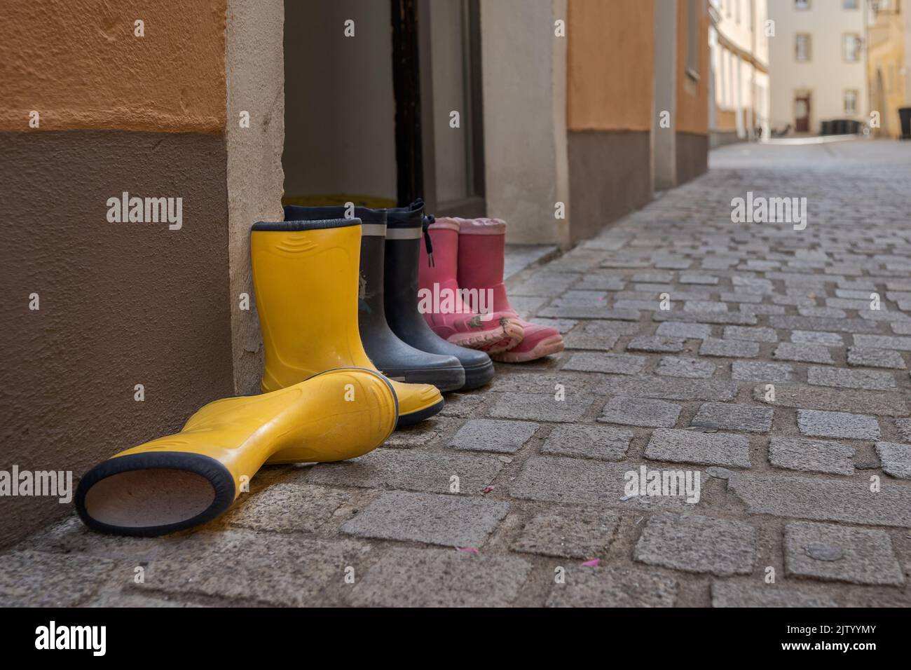 Tres pares de botas de goma para niños en diferentes colores parados en la calle al lado de la puerta delantera de la casa en un callejón urbano con adoquines Foto de stock