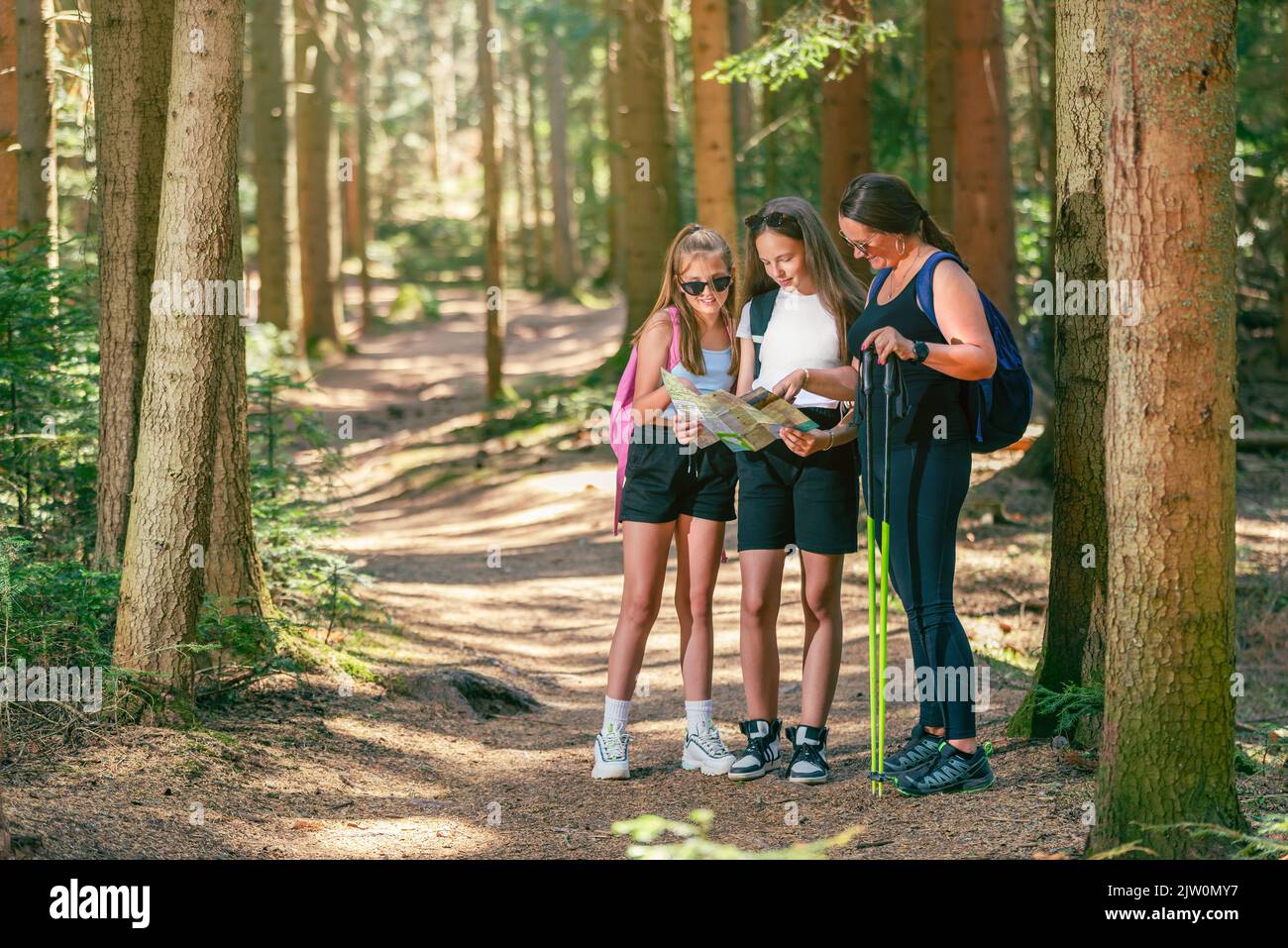 Camino del bosque con mujeres montañeras leyendo un mapa. Las chicas adolescentes sostienen un mapa mientras que una mujer sostiene postes de senderismo Foto de stock
