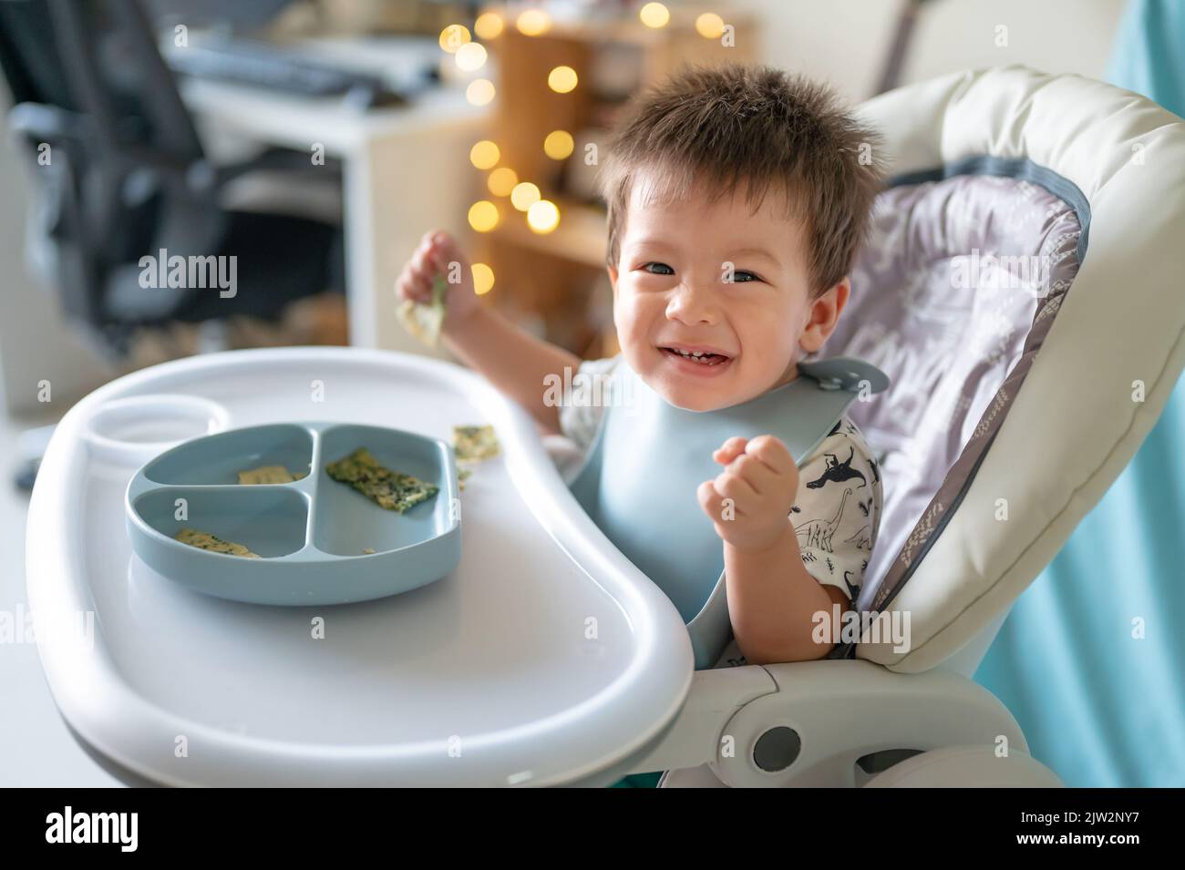 Niño comiendo solo en su silla alta en casa. Adorable bebé de un año comiendo en sus manos y comiendo en casa Foto de stock