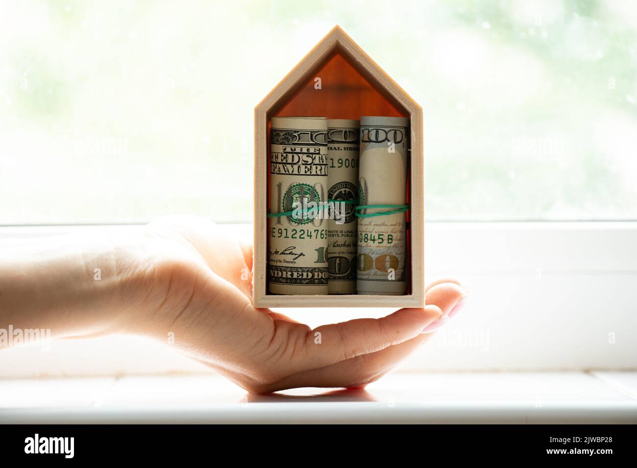 Una pequeña casa de madera con billetes de cien dólares en el interior en la ventana de la casa en manos de una niña, comprando una casa, finanzas y bienes raíces Foto de stock