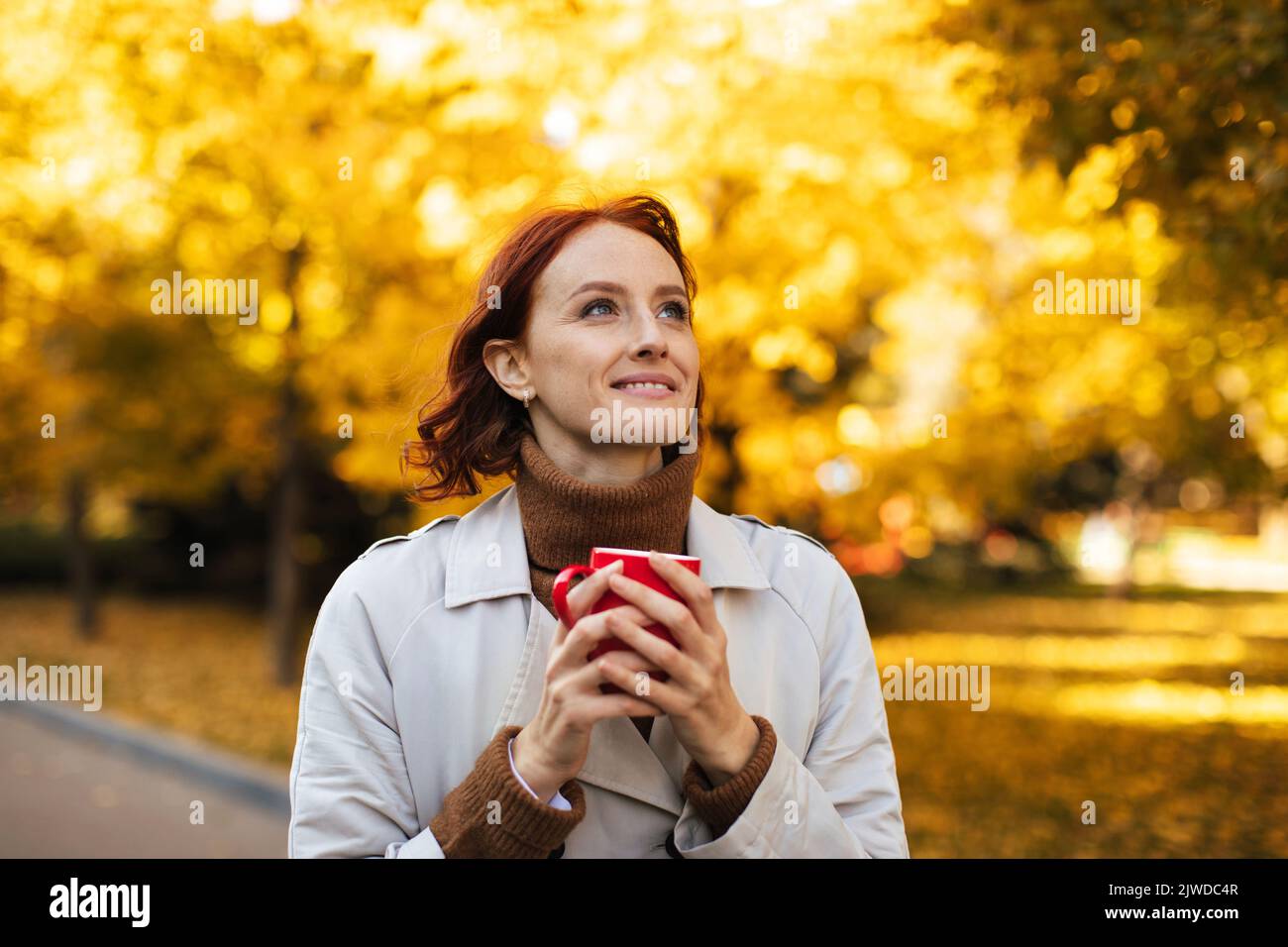 Mujer europea joven pelirroja feliz en chubasquero con taza de bebida caliente favorita, disfrutar de la libertad y el tiempo Foto de stock