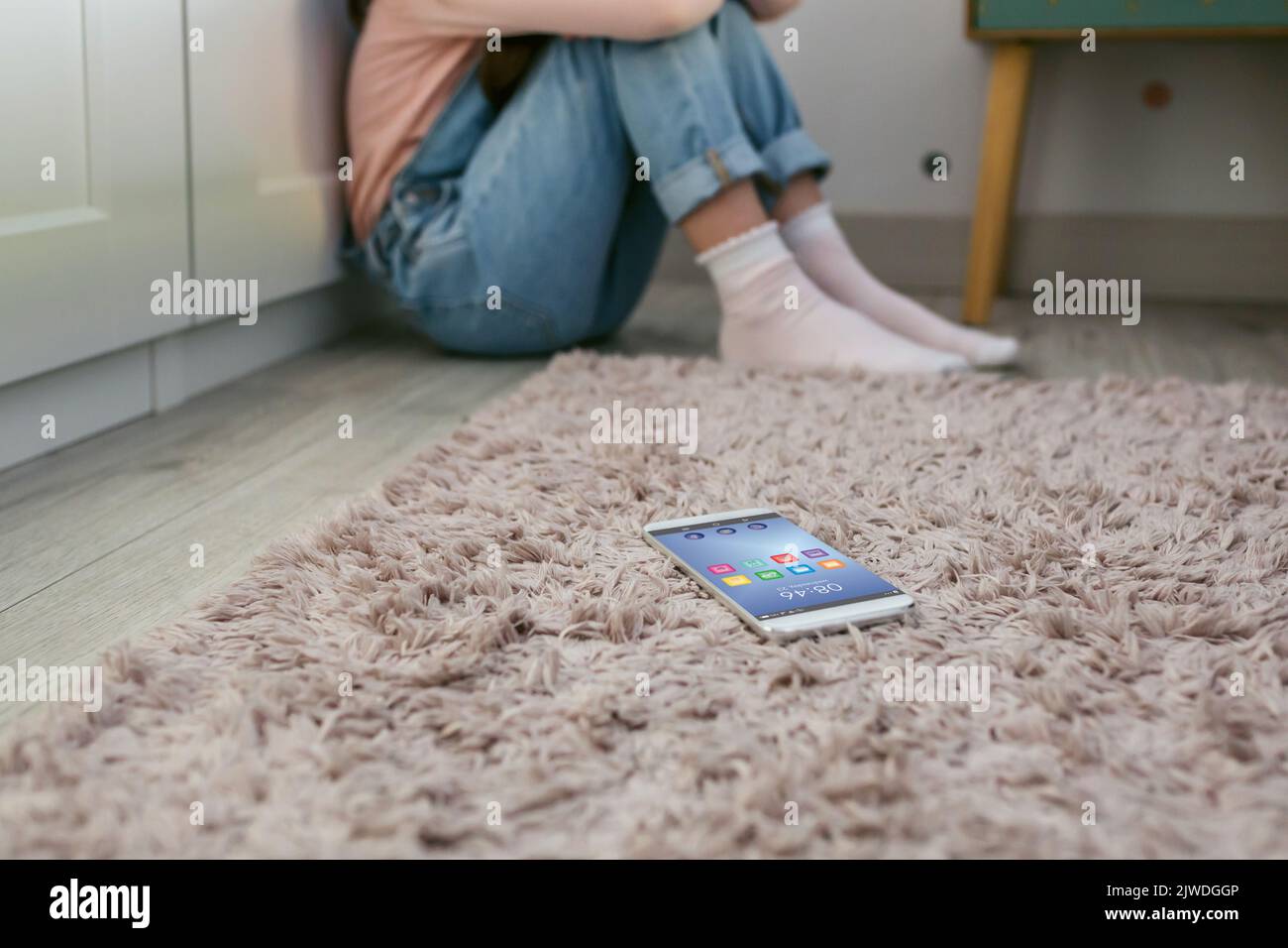 Teléfono móvil tumbado en el suelo con una niña triste irreconocible sentada detrás Foto de stock