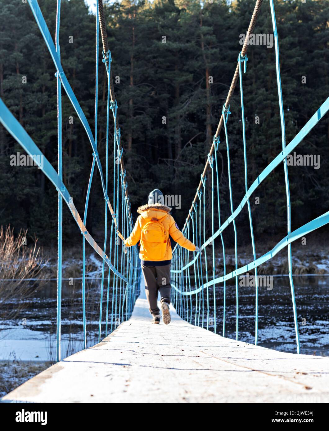Vista trasera de una mujer joven con chaqueta amarilla con mochila caminando a lo largo de un viejo puente nevado de madera a través del río en otoño e invierno contra las caminatas por el bosque Foto de stock