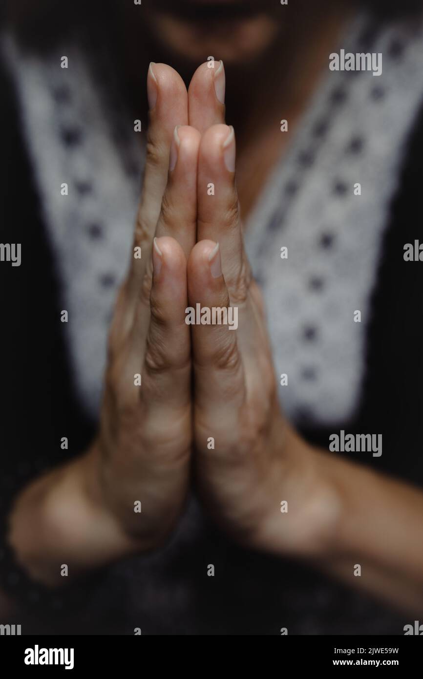Primer plano de mujer orando o meditando con sus manos juntas en namaste gesto y cuerpo borroso en el fondo en una atmósfera de humor oscuro Foto de stock