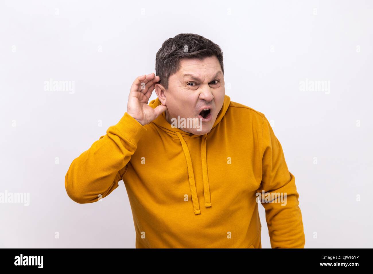 No puedo oírle. Retrato de un hombre atento y guapo de pie con la mano en el oído y que quiere oír algo, con una sudadera con capucha de estilo urbano. Estudio de interior grabado aislado sobre fondo blanco. Foto de stock