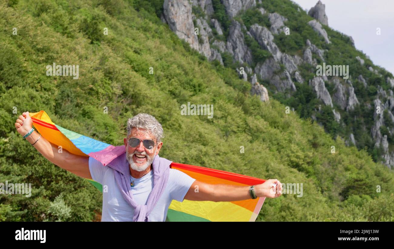 Retrato de un hombre de raza gris, anciano, caucásico, bisexualidad con barba y gafas de sol con una bandera LGBTQIA arco iris en la naturaleza. Celebra el Mes del Orgullo, el Día de la Bandera del Arco Iris, desfile gay Foto de stock