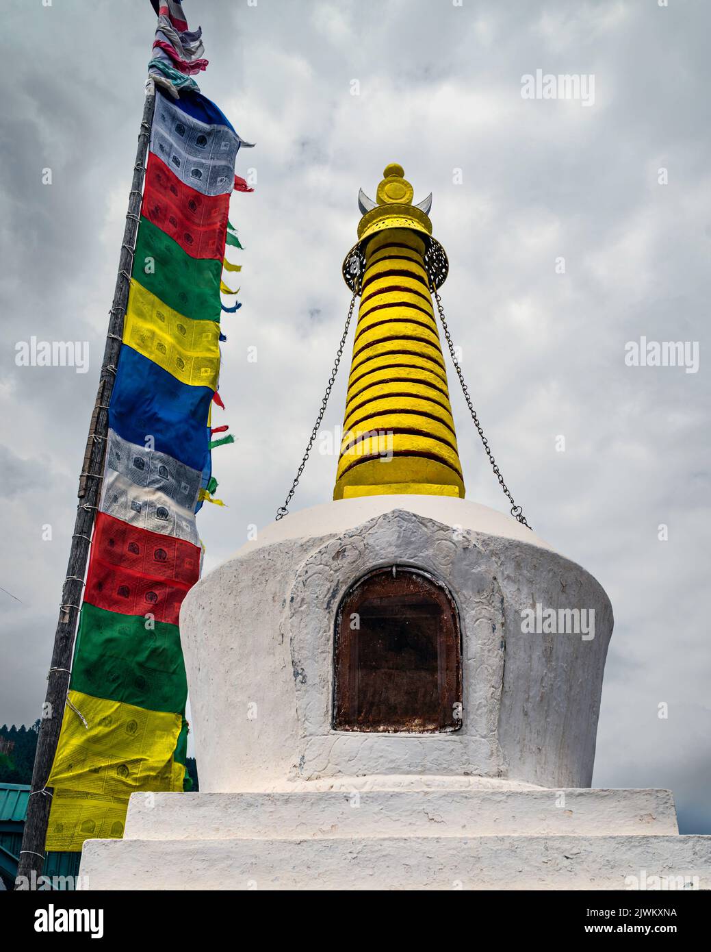 Estupa Buddist colorida flanqueada por banderas de oración de varios colores adheridas a postes de madera, todo bajo cielo nublado al aire libre en Kalpa, India. Foto de stock