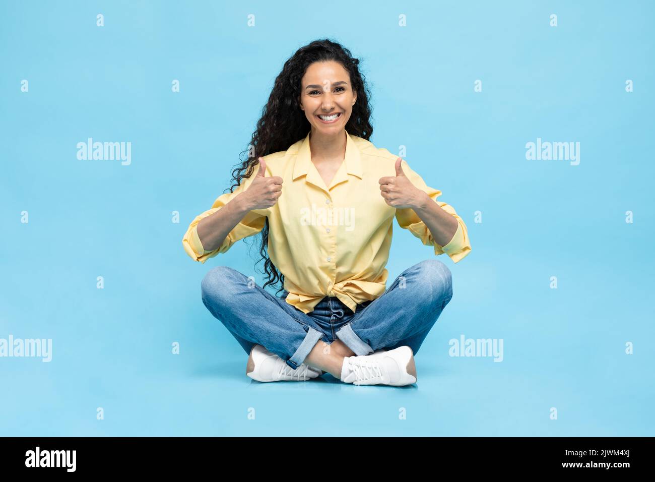 Feliz árabe femenino gesturing Thumbs arriba sentado sobre fondo azul Foto de stock
