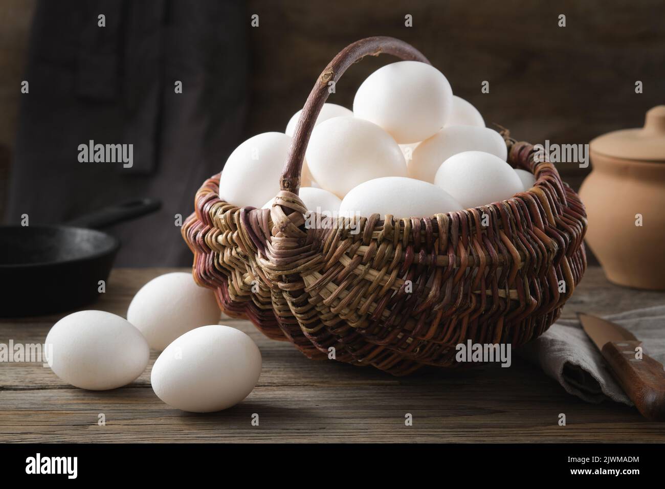 Cesta de mimbre llena de huevos de pollo blancos sobre una mesa de madera. Sartén y olla de arcilla en el fondo. Foto de stock