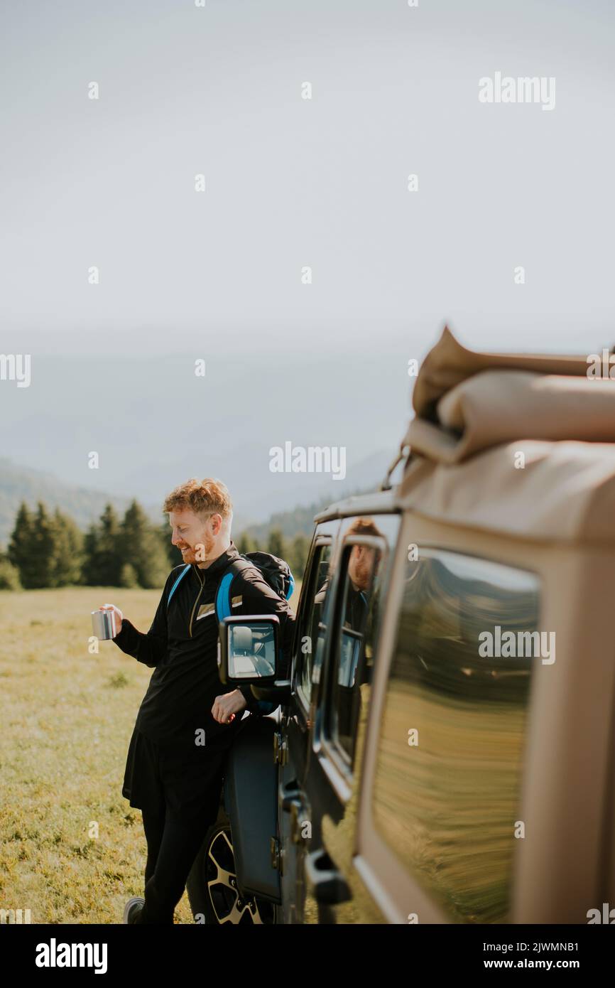Hombre joven guapo, relajándose y bebiendo café junto al capó del vehículo en el campo Foto de stock