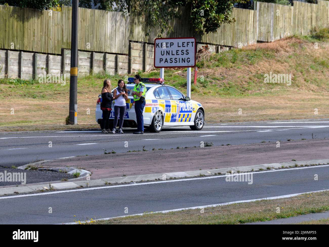 Un coche patrulla de la policía que pasa se ocupa de las dos jóvenes turistas que fueron enganche-senderismo en una carretera principal muy transitada en Taupo, cerca del centro de Nueva Zealan Foto de stock
