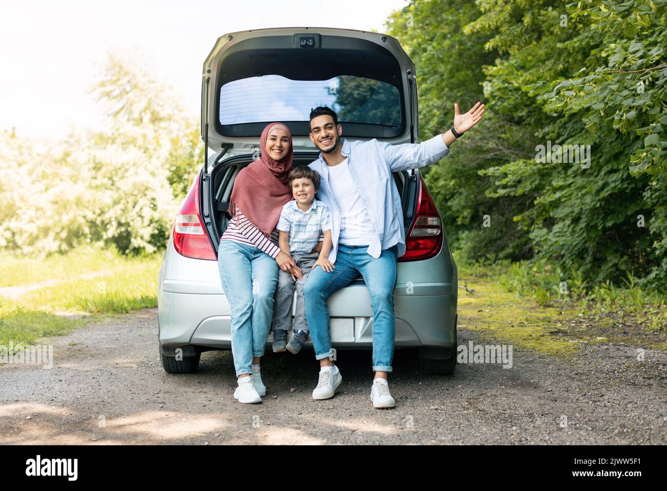 Trayecto automático positivo. Feliz milenaria árabe tipo y dama en hijab con el bebé, sentarse en el maletero del coche y gesturing Foto de stock