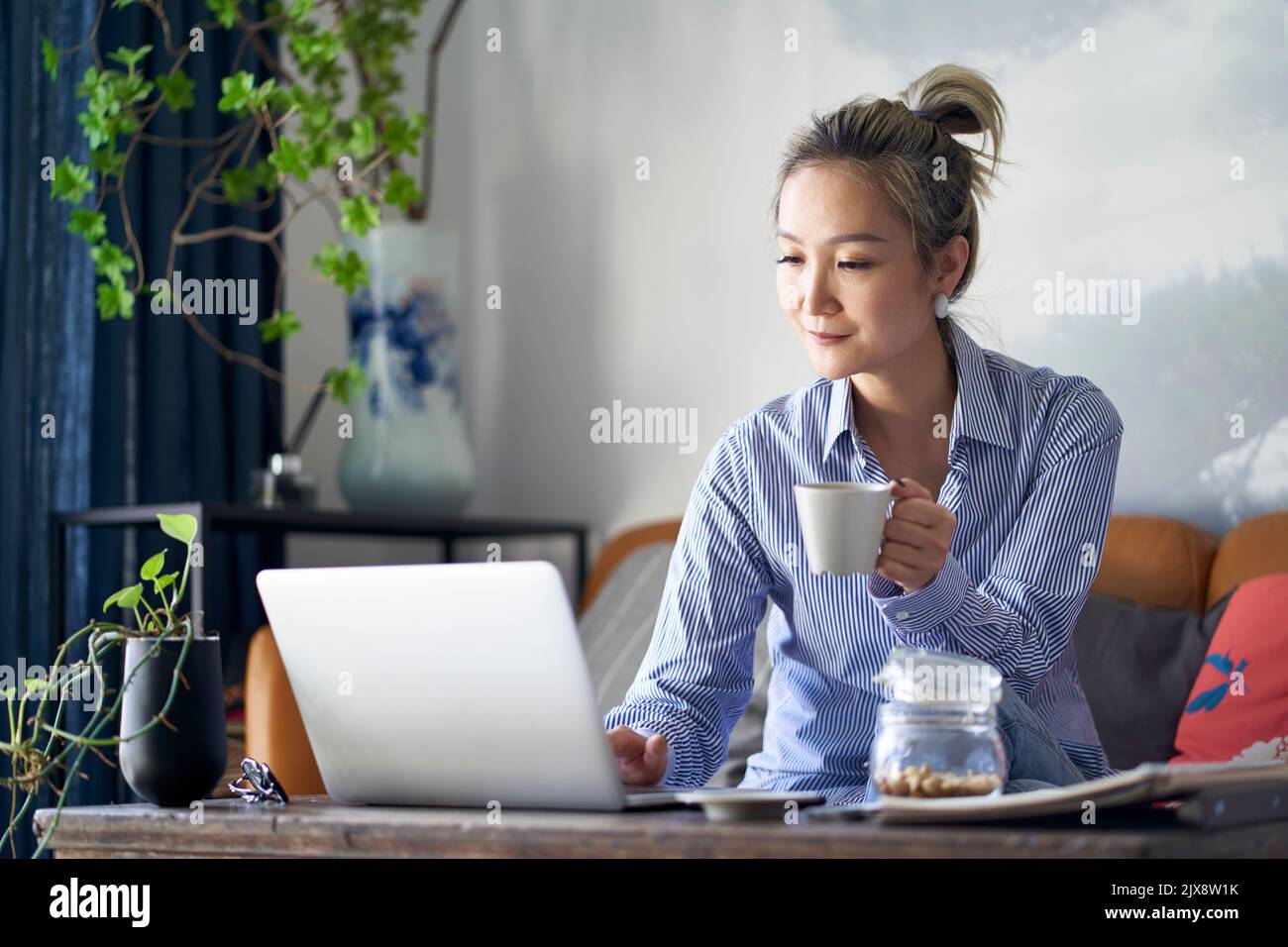 mujer asiática profesional madura que trabaja desde casa sentada en el sofá bebiendo café mientras mira la computadora portátil Foto de stock