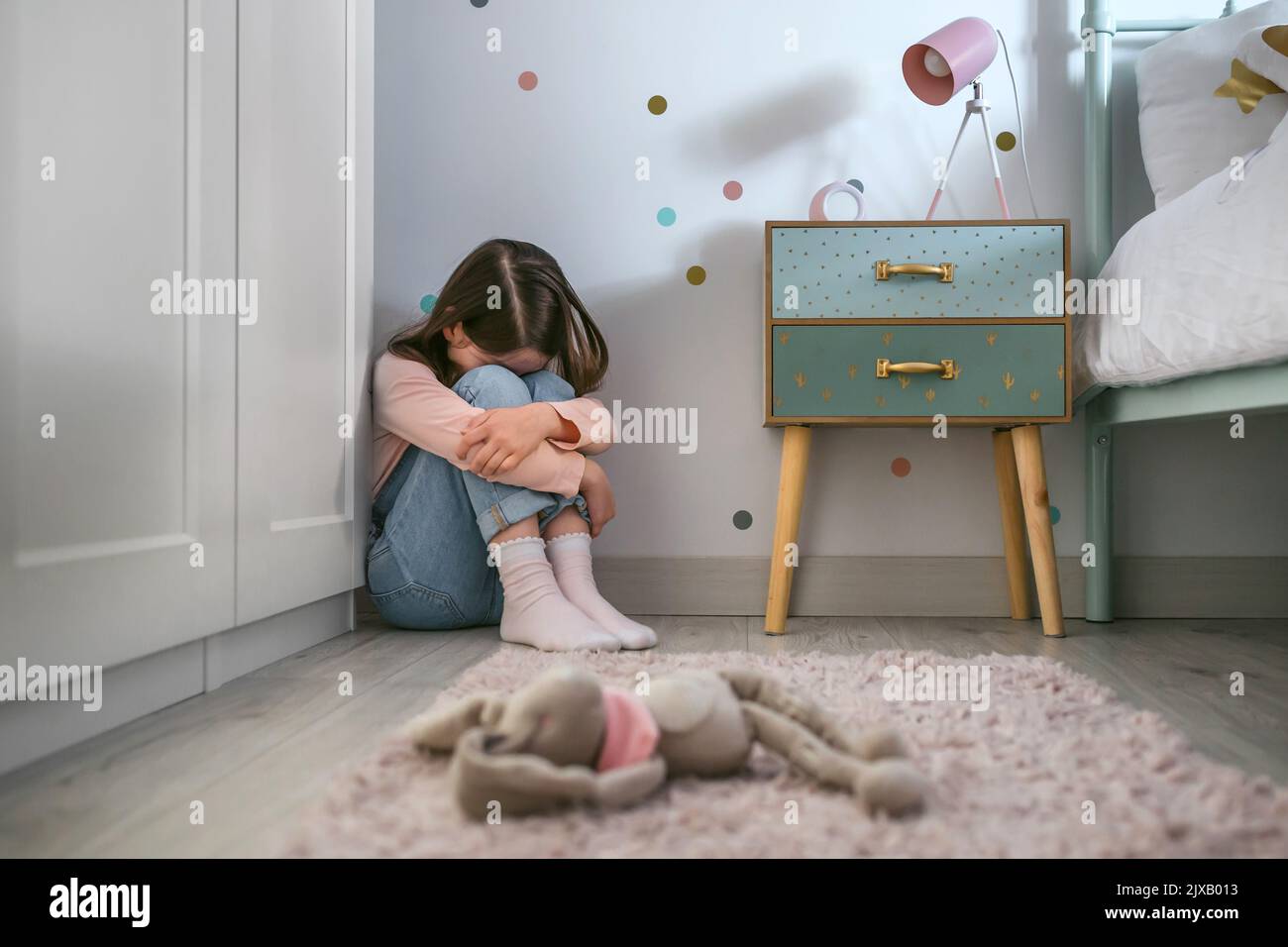 Triste niña sentada en el suelo de su dormitorio con juguete de peluche tumbado Foto de stock