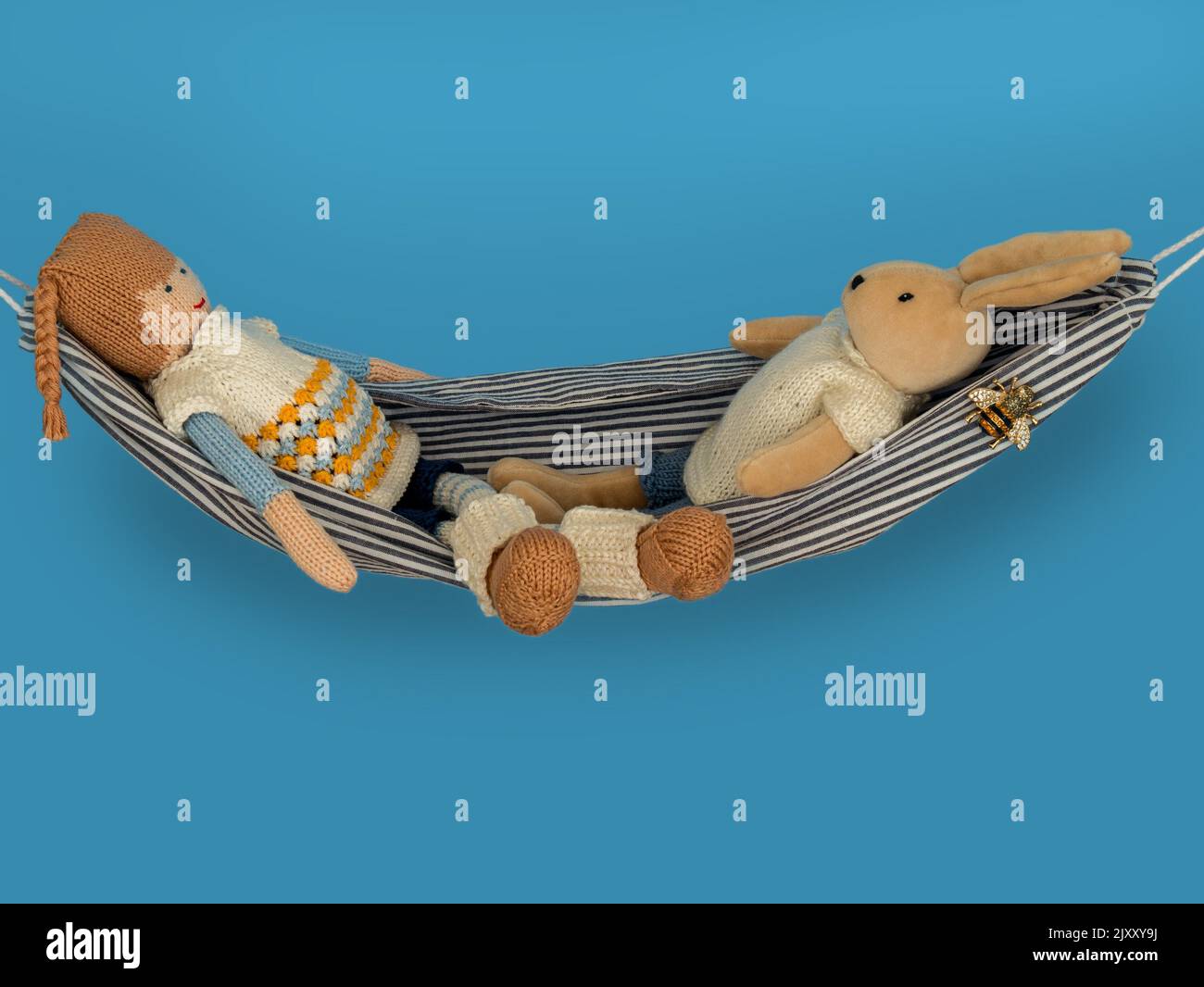 Imagen del concepto de relajación que muestra los juguetes blandos de dos niños (un conejo y una muñeca) relajándose en una hamaca frente a un fondo azul. Foto de stock