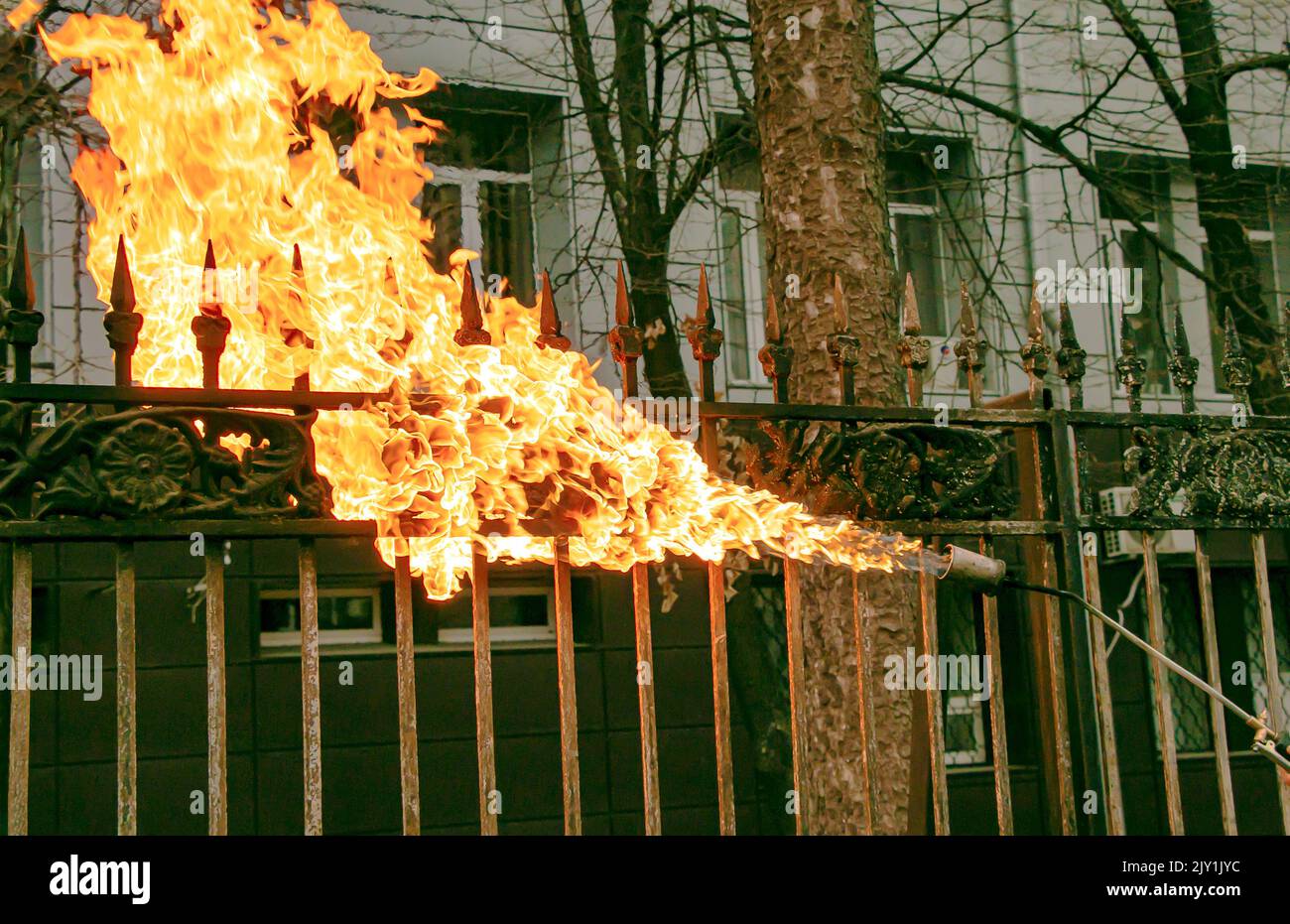 Un trabajador limpia la pintura vieja de una cerca de metal usando fuego. La mano de un hombre sostiene un quemador de gas con una llama encendida. Foto de stock